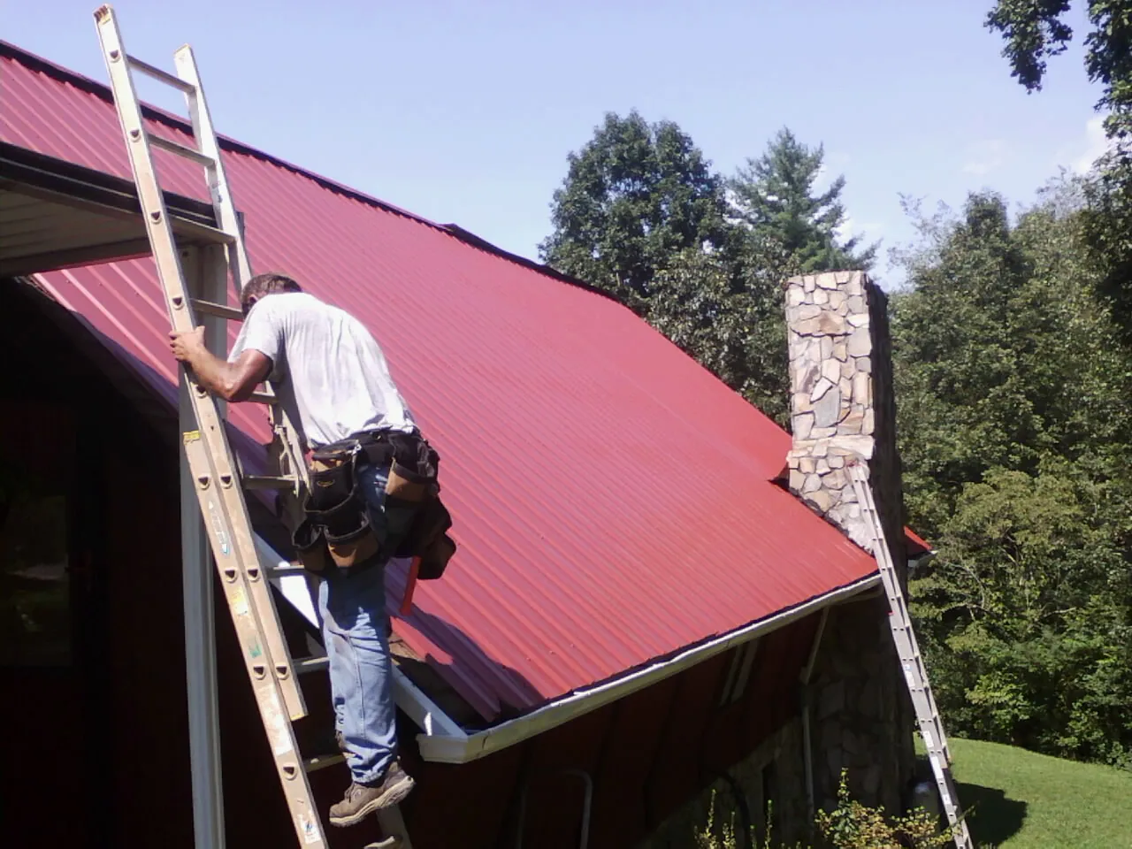 Roofer on a ladder beside a red metal roof and stone chimney — Soffit & Fascia Repair by Roofing Brothers in Castalian Springs